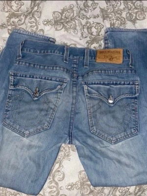 True Religion ljusblå jeans - Säljer, ett par ljusblå jeans från True Religion. Jeansen har raka ben, slitningar framtill och detaljerade knappar. Klassisk läderpatch bak.