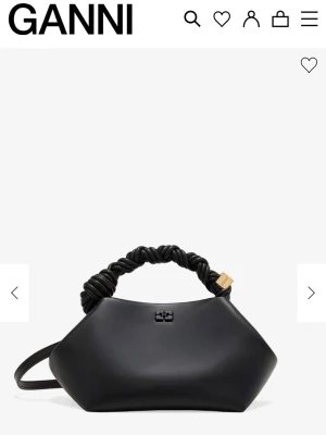 Svart handväska från Ganni - Snygg svart handväska från Ganni med unik, vriden handtagsdetalj och geometrisk form. Väskan har ett litet metallogo framtill och en axelrem. Tillverkad i slätt material som ger en modern och stilren look. Nypris: 4 295kr.