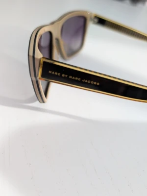 Svarta solglasögon Marc Jacobs - Solglasögon från Marc by Marc Jacobs med svart och beige båge samt gulddetaljer. Glasen är mörka och bågarna har en cool, kantig form som ger en trendig vibe. Perfekta för dig som vill sticka ut med accessoarerna. Bågarna kan enkelt justeras hos optiker om de känns för tighta bakom öronen (vanligt med glasögon).