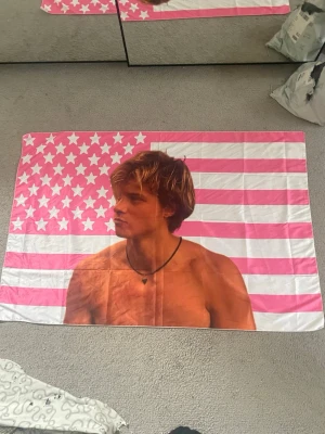 Rosa och vit flagga med JJ - En stor flagga med motiv av JJ från OuterBanks med solbrunt hår och halsband, framför en rosa och vit amerikansk flagga. Flaggan är rektangulär och gjord av tunt tyg, perfekt att ha i ditt rum på väggen eller i sängen. Jag säljer denna då jag precis hunnit sätta upp tavlor där jag tänkt den skulle va. Öppnades/hämtade flaggan 11/3 så har precis fått hem den. 