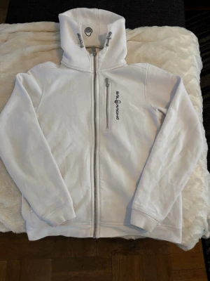 Vit hoodie från Sail Racing - Vit hoodie från Sail Racing med dragkedja framtill och en bröstficka med zip. Mjuk fleece på insidan och broderad logga på bröstet samt huvan. Snygga detaljer med svarta kontrastsömmar vid kragen. Perfekt för chill dagar.