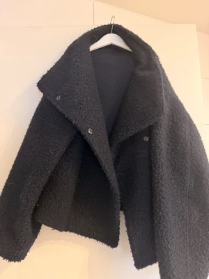 Svart teddyjacka från Gina Tricot - Mysig svart teddyjacka från Gina Tricot med bred krage och tryckknappar. Jackan har en fluffig, fårullsliknande yta och är perfekt för kyliga dagar. Oversized passform ger en avslappnad look och den är enkel att matcha till olika outfits.