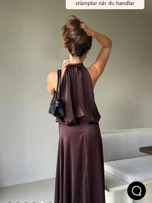 Mörkbrun satin maxiklänning - Snygg långklänning personaliserad med halterneck och öppen rygg i ett glansigt satinmaterial. Klänningen har en elegant, flytande passform och en djup mörkbrun ton som ger en lyxig känsla. Perfekt för dig som vill sticka ut med en stilren look.