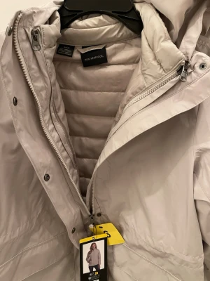 Ljusgrå regnjacka från Weatherproof - Stilren ljusgrå regnjacka från Weatherproof med huva, dragkedja och tryckknappar. Jackan har två stora fickor framtill och quiltat innerfoder för extra komfort. Perfekt för regniga dagar och enkel att matcha med olika outfits.