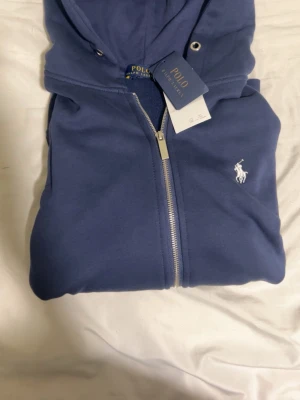Blå zip hoodie från Polo Ralph Lauren - Snygg blå hoodie från Polo Ralph Lauren med dragkedja framtill och klassisk vit logga broderad på bröstet. Hoodien har huva med dragsko och två fickor framtill. Tillverkad i mjukt bomullsmaterial för skön känsla. Perfekt för en avslappnad och stilren look.