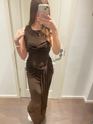 Brun långklänning från Boohoo - Snygg brun långklänning från Boohoo med knyt-detaljer i midjan och över bysten. Klänningen är ärmlös och har en figurnära passform som framhäver siluetten. Tillverkad i ett mjukt och stretchigt syntetmaterial.