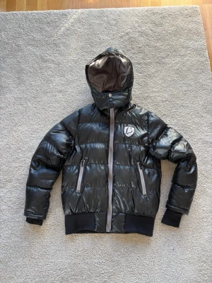 Svart pufferjacka - Snygg svart pufferjacka från Sexy by Sweden med glansig finish, huva och två dragkedjefickor framtill. Jackan har ribbade muddar vid ärmslut och nederkant samt en tydlig logga på bröstet. 