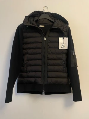 Svart pufferjacka/Cardigan  från Moncler - Snygg svart pufferjacka från Moncler med quiltad front och huva. Jackan har stickade ärmar och rygg, dragkedja framtill och fickor med knappar. Märkeslapp på bröstet och ärmficka med dragkedja. Perfekt för kyliga dagar.