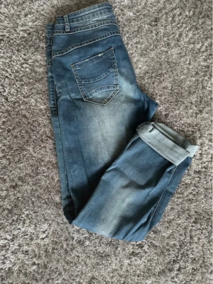 Blå jeans med uppvikta ben - Säljer ett par blå jeans med klassisk femficksdesign och uppvikta ben. Jeansen har en snygg tvätt med ljusa partier och är tillverkade i denim. Passformen är rak och de har dragkedja och knapp framtill.