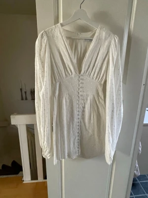 Kortklänning från Adoore - Supersöt vit kortklänning från Adoore med diskreta prickar, v-ringning och långa puffärmar. Klänningen har dekorativa knappar framtill och lätt markerad midja. Den är bara använd en gång🥰