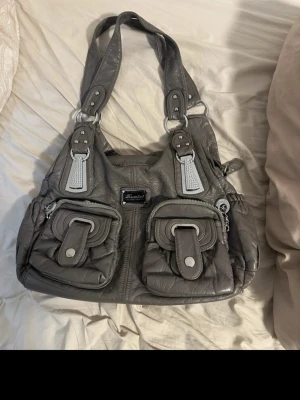 Grå handväska med dubbla fickor - Snygg grå handväska från Xhilaration i konstläder med två stora framfickor med dragkedja och spännen. Väskan har breda axelband med silverfärgade detaljer och en rektangulär form. Perfekt för dig som gillar praktiska och trendiga accessoarer.