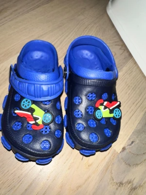 Blå sandaler med motorcykeldekor - Säljer ett par coola sandaler i blått och mörkblått med roliga motorcykeldekorationer framtill. Skorna har justerbar hälrem och är tillverkade i lätt plastmaterial med ventilerande hål. Perfekta för sommaren och riktigt bekväma att ha på sig.