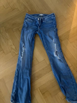 Blå slitna bootcut jeans - Säljer ett par blå bootcut jeans med slitningar och hål på båda benen. Jeansen har klassisk femficksdesign och låg midja. Perfekta för en avslappnad och trendig look.