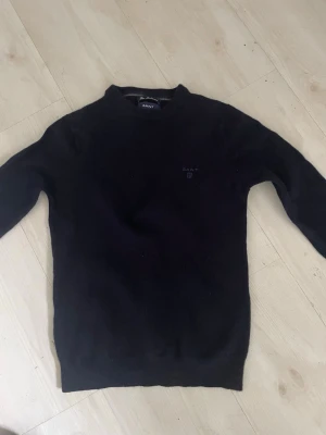 Svart stickad tröja från Gant - Klassisk svart stickad tröja från Gant med rund halsringning och diskret broderad logga på bröstet. Tröjan har långa ärmar och ribbade muddar vid ärmslut och nederkant. Perfekt basplagg för en stilren look.