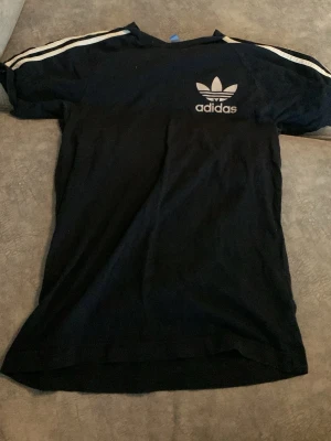 Svart Adidas t-shirt med vita ränder - Klassisk svart t-shirt från Adidas med vita ränder längs axlarna och det ikoniska Adidas Trefoil-loggan tryckt på bröstet. T-shirten har korta ärmar och är gjord i mjuk bomull för en skön känsla. Perfekt till streetwear-stilen.