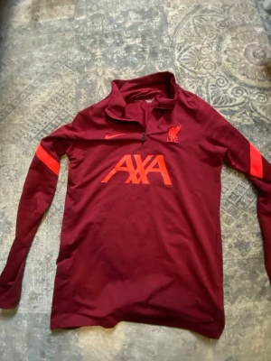 Liverpool Nike Dri-FIT träningströja - Snygg vinröd långärmad träningströja från Nike med Liverpool FC-logga och AXA-tryck på bröstet. Tröjan har halv dragkedja, Dri-FIT-material och röda detaljer på ärmarna. Perfekt för fotbollsträning eller sportiga dagar.