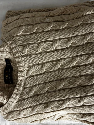 Beige kabelstickad tröja Polo Ralph Lauren - Kabelstickad tröja i beige från Polo Ralph Lauren. Klassisk rund halsringning och ribbade muddar. Tillverkad i mjukt material med tidlös design och diskret logga på insidan av kragen. Perfekt för lager på lager och enkel att matcha.