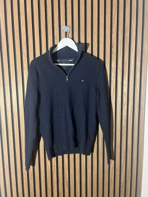 Mörkblå halvzip från Tommy Hilfiger - Snygg mörkblå stickad tröja från Tommy Hilfiger i extra fin ull. Tröjan har halv dragkedja, liten logga på bröstet och ribbade muddar. Perfekt till lager på lager och enkel att matcha med jeans eller chinos. Storleken är M men sitter som en S.