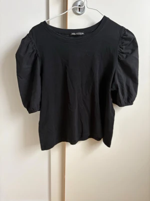 Svart topp med puffärm från Zara - Svart topp från Zara med puffiga korta ärmar och rund halsringning. Toppen är gjord i mjuk bomull och har en enkel, stilren design som passar till mycket. Perfekt för dig som gillar en trendig men basic look. Tröjan är sparsamt använd så mycket fint skick!! 