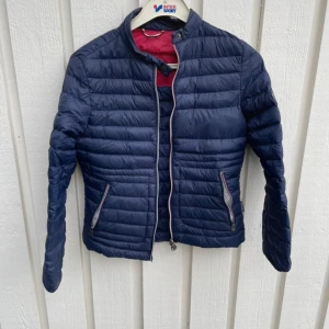 Mör them blå pufferjacka Key West - Snygg mörkblå p djupufferjacka från Key West expend med quiltad design och två dragkedjeförsed specda fickor. Jackan har en hög krage och kontrastf Resolve ärgad insida i rött. Perfekt för kalla dagar och ger en sportig look.