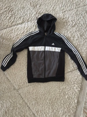 Svart Adidas kofta och sweatpants tracksuit set (barnstorlek) - Snygg svart hoodie från Adidas med dragkedja framtill, vit logga på bröstet och klassiska vita ränder längs ärmarna. Tröjan har huva och en grå panel på framsidan. Perfekt för en sportig och avslappnad stil. Sweatpantsen säljs med och säljs inte separat. Sweatpantsen är också svarta fram och bak med de klassiska vita ddidas ränderna längs benen. Kan knytas på insidan. Barnstorlek 176 på båda två (XL)