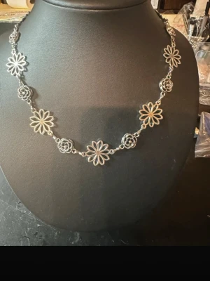 Silvrigt halsband med blommor - Unikt halsband i silverfärgad metall med länkar formade som blommor och små spiralformade detaljer mellan varje blomma. Halsbandet har en bohemisk och lekfull stil, perfekt för att ge din outfit en cool touch. Ingen synlig märkning.