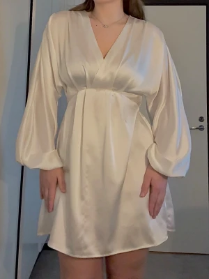 Vit klänning i satin / Gina Tricot  - Kort vit klänning i glansigt satinmaterial med v-ringning och långa, lösa ärmar. Klänningen har omlott-look framtill och markerad midja som ger en smickrande siluett. Perfekt för dig som gillar en elegant och stilren vibe.