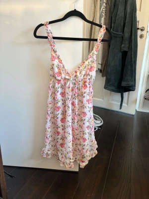 Blommigt vitt nattlinne med volang - Supersött vitt nattlinne med rosa och grönt bl proven blommönster. Har volangkant nertill och längs axelbanden samt en liten rosett framtill. Axelbanden är breda och nattlinnet har en luftig, flowy passform. Perfekt för mys phone kvällar hemma. Från hm i storlek M, ganska kort och lite genomskinligt!🌟🌟