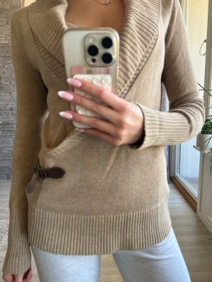 Beige omlottstickad tröja med spänne - Säljer en beige stickad omlott-tröja med bred krage och snyggt brunt spänne på sidan. Super snygg sitter som en s 💕