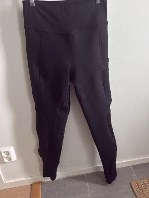 Svarta ridleggings med silikon - Säljer ett par svarta ridleggings med hög midja och silikonmönster på insidan av benen och bakdelen för extra grepp. Leggingsen har praktiska fickor på sidorna och är tillverkade i ett stretchigt material som ger bra rörelsefrihet. Fodrade ridbyxor 
