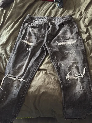 Grå slitna jeans med hål - Säljer ett par svarta jeans från supply & demand med tydliga slitningar och hål på både fram- och baksida. Jeansen har en relaxed fit och klassisk femficksmodell. Materialet är denim i bomull och färgen är tvättad svart med vita detaljer och slitna kanter.
