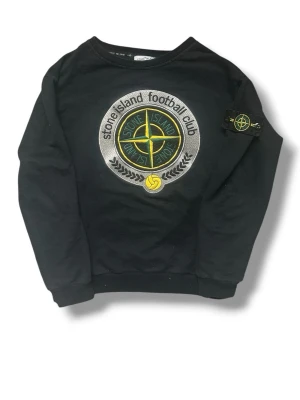 Svart Stone Island sweatshirt S - Svart sweatshirt från Stone Island med stort broderat logomärke på bröstet och klassisk patch på vänster ärm. Tröjan har rund hals, ribbade muddar och är tillverkad i mjuk bomull. Perfekt för dig som gillar streetwear och sportig stil. (Barnstorlek) så passar xs
