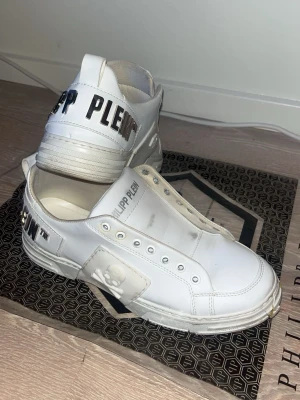 Vita Philipp Plein sneakers - Snygga vita sneakers från Philipp Plein med metallic-logga på hälen och präglad dödskalle på sidan. Skorna är låga och har detaljer i vitt läder samt silverfärgade nitar baktill. Perfekta för dig som gillar streetstyle och exklusiva märken. OBS SKORNA HAR ETT LAGAT HÅL PÅ FRAMMSIDAN!!!