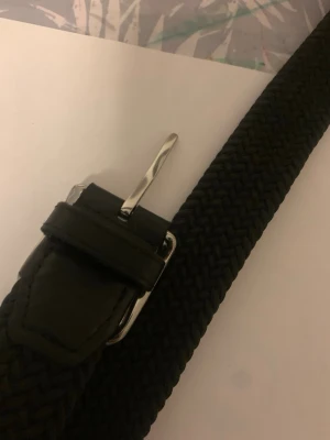 Svart flätat bälte med silverfärgat spänne - Stilrent svart flätat bälte med ett snyggt silverfärgat spänne. Bältet har en klassisk design och är gjort i ett syntetmaterial med detaljer i skinnimitation. Perfekt accessoar för att lyfta din outfit.