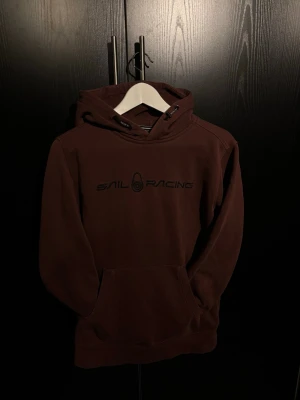 Vinröd/mörkröd hoodie från Sailracing - Vinröd/ mörkröd hoodie från Sail Racing med stor ficka framtill och justerbar huva med snören. Tryckt logga på bröstet i svart. Perfekt för en avslappnad och sportig stil. Mjuk bomullskänsla och ribbade muddar vid ärmslut och nederkant.