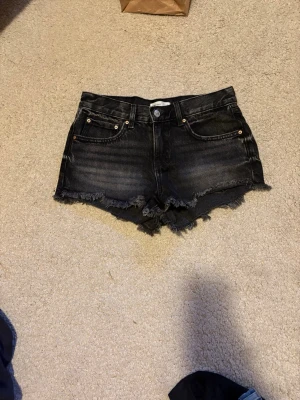 Svarta jeansshorts med fransig kant - Säljer ett par svarta jeansshorts med fransig, rå kant och klassisk femficksdesign. Shortsen har knapp och dragkedja framtill samt bälteshällor. Perfekta för dig som gillar en avslappnad och trendig stil.