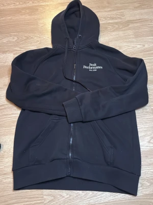 Svart hoodie från Peak Performance - Svart hoodie med dragkedja från Peak Performance. Broderad logga på bröstet och  huva med dragsko. Två fickor framtill och ribbade muddar vid ärmar och nederkant. I storlek M.