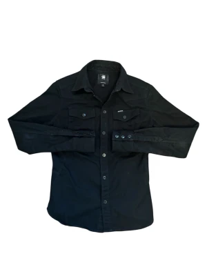Svart skjorta från G-Star RAW - Stilren svart skjorta från G-Star RAW med klassisk krage, långa ärmar och knäppning framtill. Två bröstfickor med knappar ger en cool utility-vibe. Perfekt till jeans eller chinos för en avslappnad men snygg look.