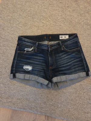 Mörkblå jeansshorts  - Säljer ett par mörkblå jeansshorts från Crocker med slitna detaljer och upprullade benslut. Klassisk femficksmodell med knapp och dragkedja framtill. Perfekta för sommaren och ger en cool, avslappnad look.