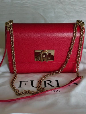 Rosa axelväska från Furla - Snygg röd axelväska från Furla i läder med guldfärgad kedjerem och elegant låsdetalj framtill. Stilren och kompakt design som ger en lyxig känsla. Perfekt accessoar för att lyfta din outfit.