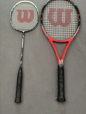 Wilson tennis- och badmintonracket - Två racketar från Wilson, en för tennis i rött och svart med stort huvud och en för badminton i svart och blått. Båda har svart grepp och tydlig Wilson-logga på strängarna. Perfekt för dig som gillar racketsport och vill ha både tennis- och badmintonracket.