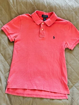 Rosa pikétröja från Polo Ralph Lauren - Klassisk rosa pikétröja från Polo Ralph Lauren med korta ärmar och krage. Tröjan har två knappar framtill och den ikoniska mörkblå logotypen broderad på bröstet. Tillverkad i mjuk bomull för en bekväm känsla.
