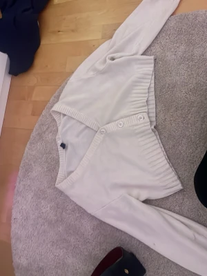 Vit cropped kofta med knappar - Säljer en vit cropped kofta med långa ärmar och tre knappar framtill. Koftan har bred ribbstickad kant vid ärmslut och nederkant, vilket ger en snygg och modern look. Perfekt att slänga över en topp för en enkel men stilren outfit.