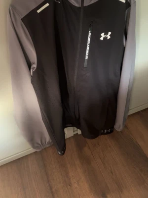 Under Armour svart/grå vindsjacka - Snygg vindjacka jacka från Under Armour i svart och grått med huva och dragkedja framtill. Jackan har logotyp på bröstet och detaljerade ventilationshål på sidorna. Materialet är lätt och funktionellt, perfekt för sport och löpning.