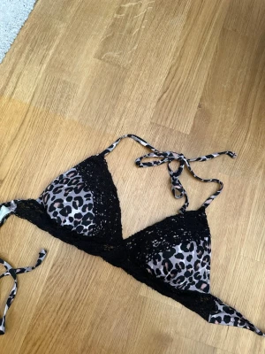 Leopardmönstrad spets-bikini med knyt - Snygg bikini med leopardmönster i beige, svart och brunt. Bikinin har tunna axelband och knyt i nacken samt spetsdetaljer i svart runt kuporna. 