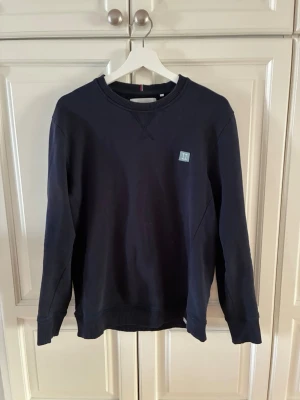 Mörkblå sweatshirt från Les Deux - Mörkblå sweatshirt med rund halsringning och ribbade muddar vid ärmslut och nederkant. Liten logotyp-patch på bröstet. Klassisk och enkel design i mjukt material.