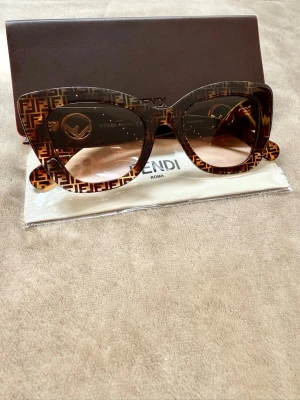 Fendi solglasögon med FF-mönster - Snygga solglasögon från Fendi med klassiskt FF-mönster i brunt och guld på bågarna. Modellen har breda, fyrkantiga bågar och tonade linser. Materialet är högkvalitativ plast och logotypen syns tydligt på sidorna. Perfekt för dig som vill sticka ut med en lyxig accessoar.