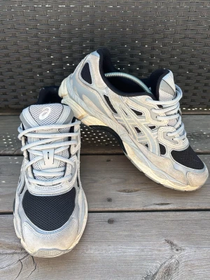 Asics sneakers i grått och svart - Snygga Asics Nyc gel sneakers med grå och svarta meshpaneler, vita detaljer och klassisk logga på plösen. Skorna har en chunky sula och sportig design, perfekta för en streetwear-look. Materialet är en mix av mesh och syntet för bra andningsförmåga.