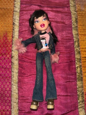 Bratz Yasmin – original outfit – Y2K / tidigt 2000-tal - Original Bratz Yasmin från tidigt 2000-tal med komplett Y2K-inspirerad outfit. Dockan bär mörka bootcut-jeans, jeansjacka med rosa fuskpälsdetaljer, rosa topp samt platåskor – en klassisk Bratz-look från den tidiga MGA-eran.  Dockan har långt mörkt hår med lugg och välbevarat smink i typisk Y2K-stil. Ansiktet är fint bevarat med tydliga läppar och ögonmakeup. Outfiten matchar den ikoniska Bratz-estetiken som är populär bland samlare idag.  Kläderna är hela och dockan är i mycket fint begagnat sk