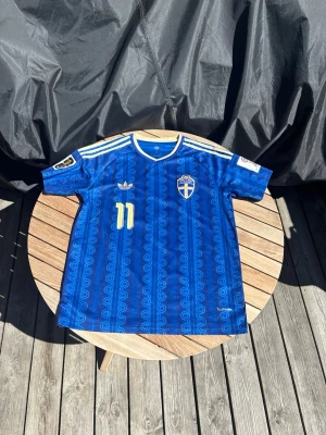 Blå Sverige fotbollströja Adidas - Säljer en blå Sverige fotbollströja från Adidas med gult nummer 11, guldfärgade detaljer och klassiska Adidas-ränder på axlarna. Tröjan har FIFA-märke på ärmen och ett mönster över hela tyget. Perfekt för dig som älskar fotboll och vill sticka ut på läktaren.